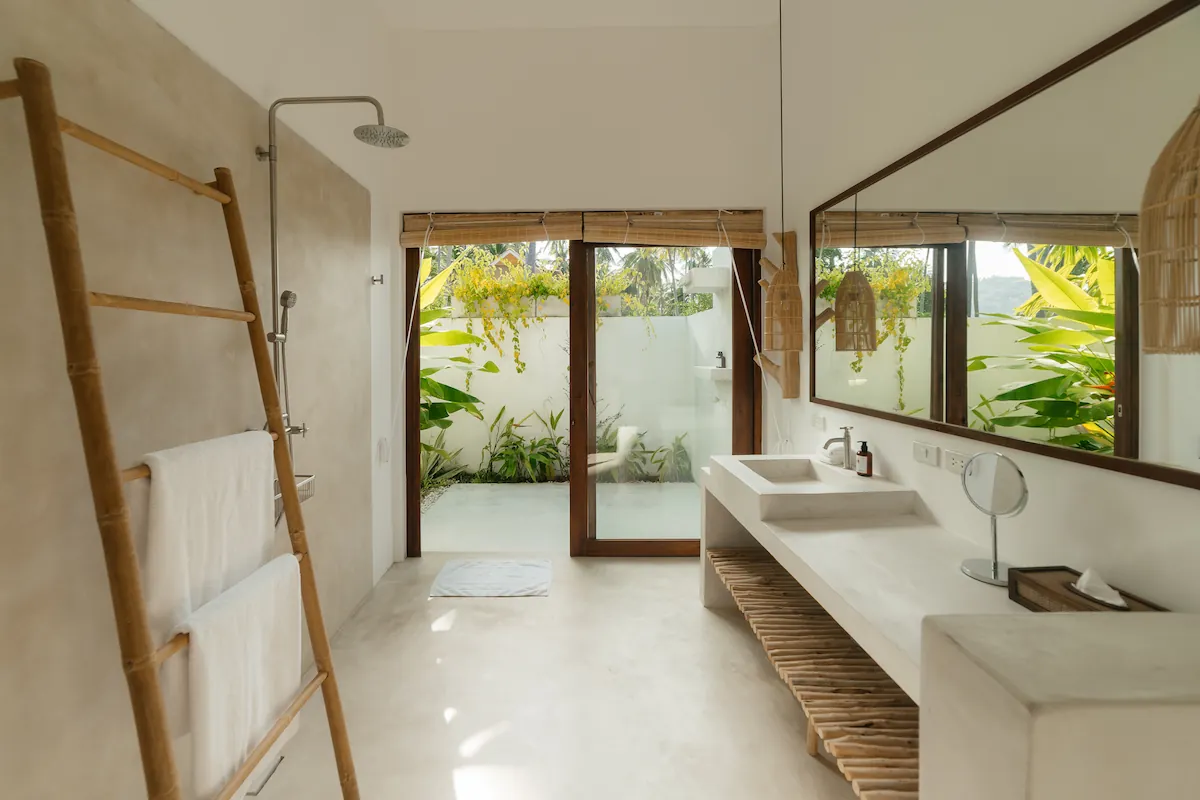Minimalist en-suite bathroom with bamboo towel ladder, concrete vanity, and glass doors to outdoor shower garden at Villa Aditi, Koh Samui